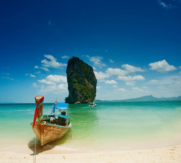 A traditional wooden long-tail boat on a pristine beach in Thailand, with turquoise waters, a towering limestone cliff, and a bright blue sky.